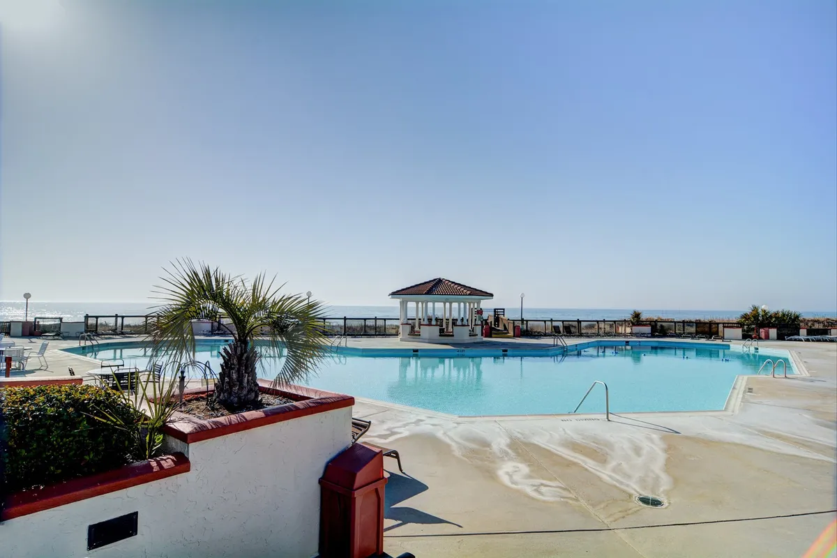 An inviting exterior scene showcasing a large swimming pool surrounded by palm trees and a poolside gazebo. The setting exudes a peaceful vibe, ideal for relaxing, swimming, and hosting poolside gatherings with friends.