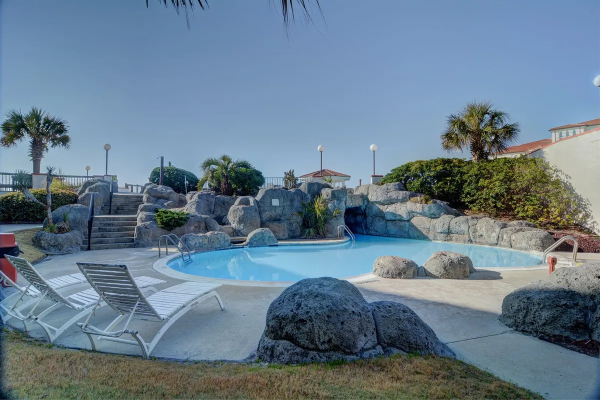 An inviting exterior scene with unique rock formations, a sparkling swimming pool, lounge chairs, and swaying palm trees. The setting is perfect for swimming, relaxing, or gathering with friends after an active day.