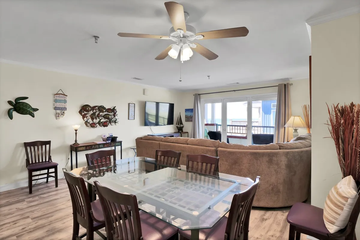 The image showcases an inviting open-concept living and dining area featuring a cozy sofa, a dining table, a television, and sliding glass doors that lead directly out to a private balcony. Decorative wall art adds personality, and the arrangement encourages both relaxation and social gatherings.