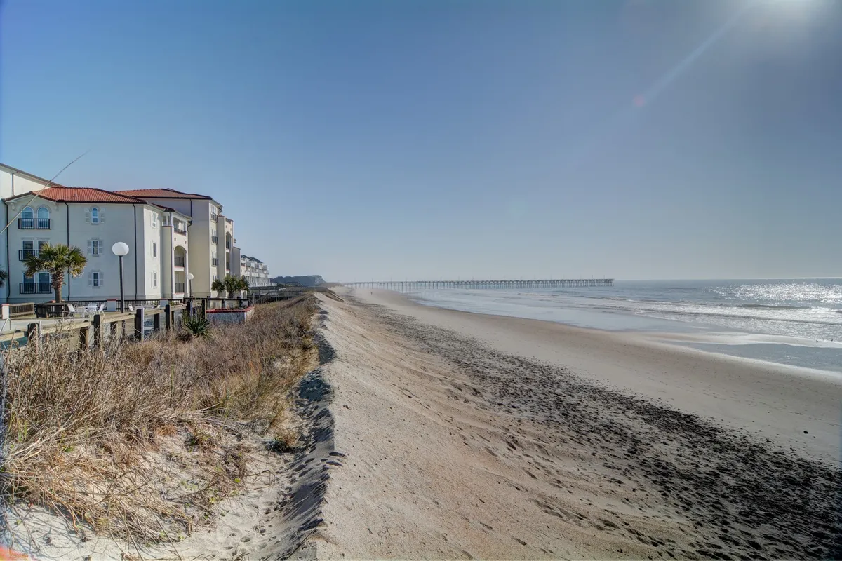 This scene features sandy beachfront properties with the iconic pier extending into the ocean. Families and children can be seen walking and playing on the beach, with the sun casting a warm, welcoming glow across the sand and surf.