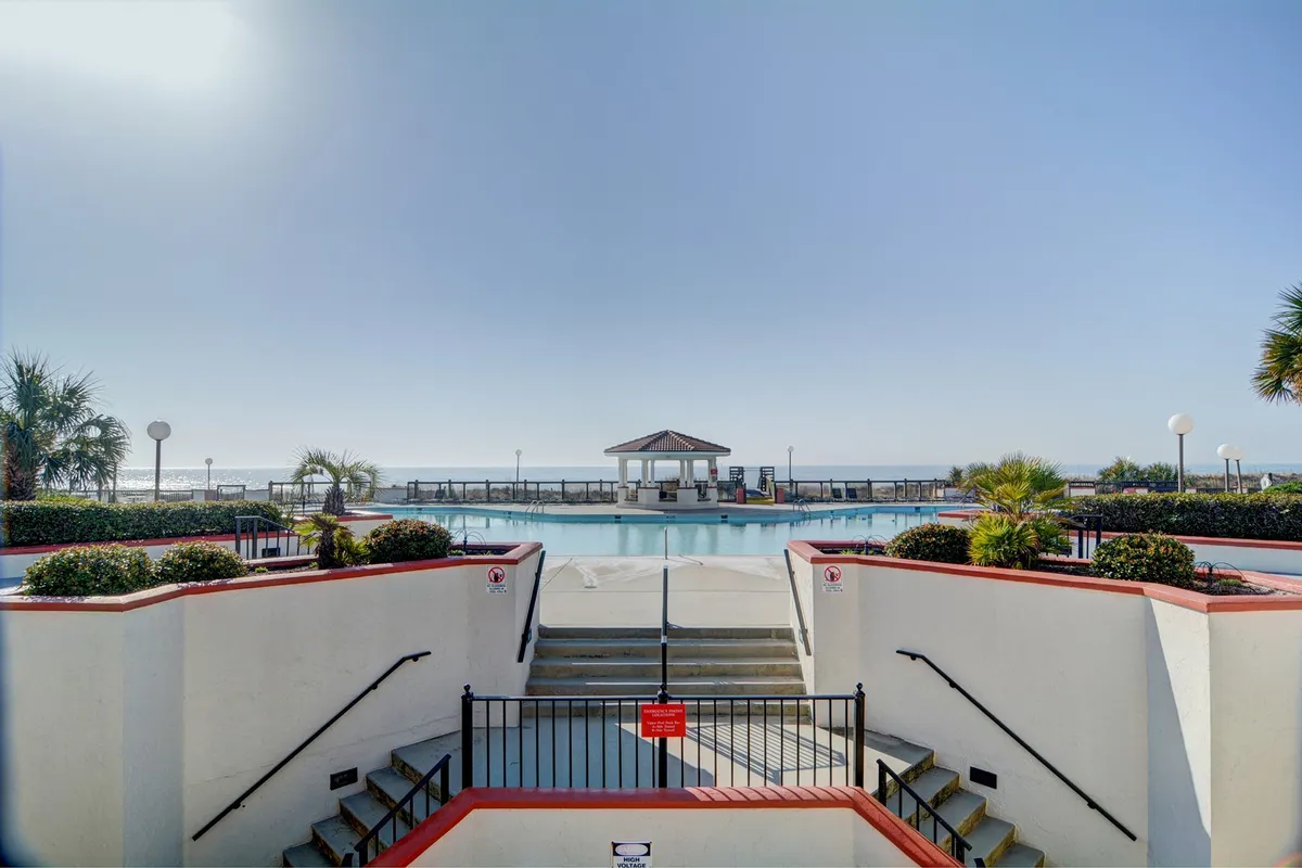 An expansive resort-style swimming pool is surrounded by palm trees, a gazebo, and sun loungers, with the ocean visible beyond. The scene exudes relaxation, with people enjoying the tranquil poolside and coastal ambiance.