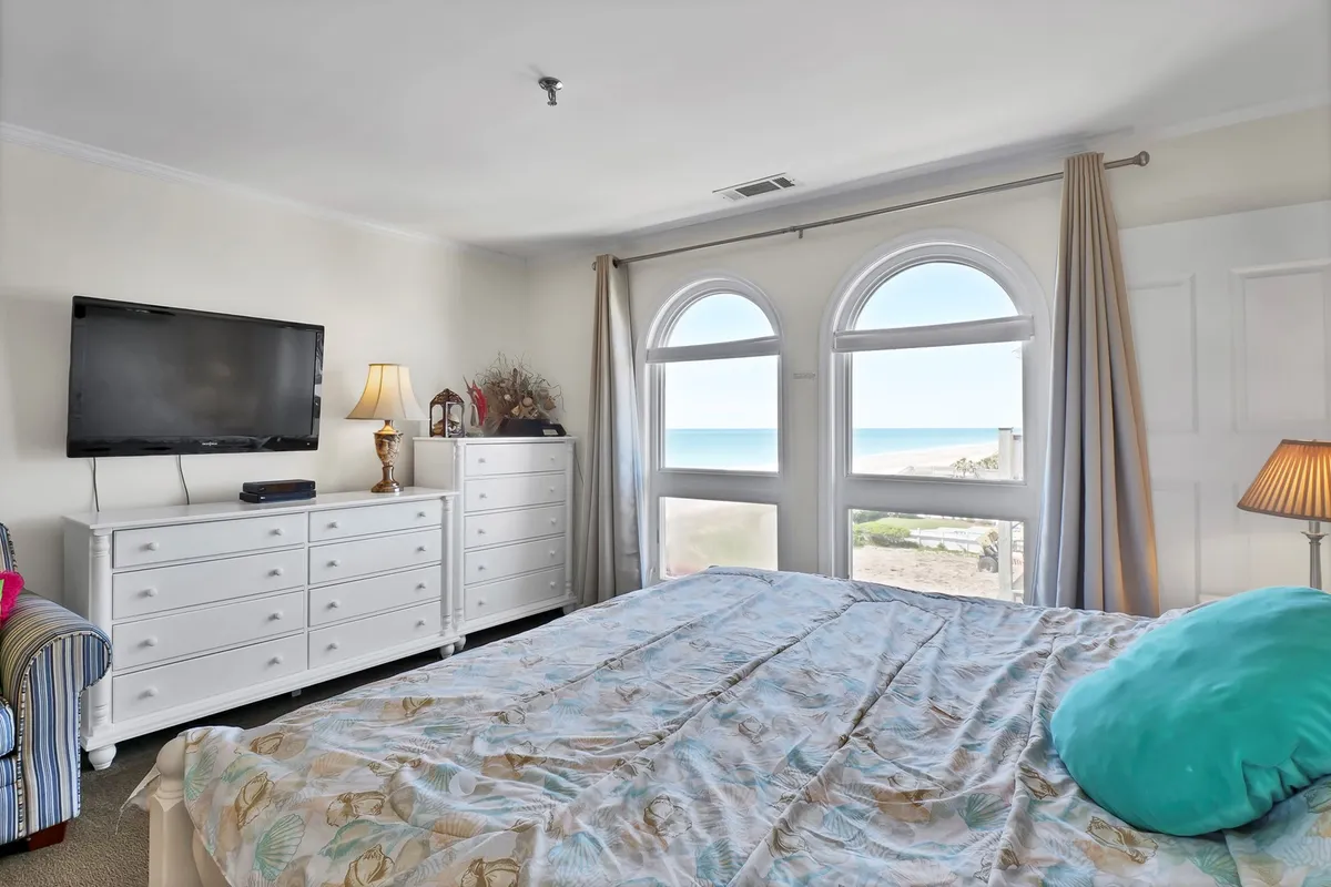 This image showcases a tranquil, inviting bedroom with a comfortable bed adorned with decorative pillows, a dressing table with a lamp, and large windows offering an ocean view. The room feels private, airy, and perfect for relaxation.