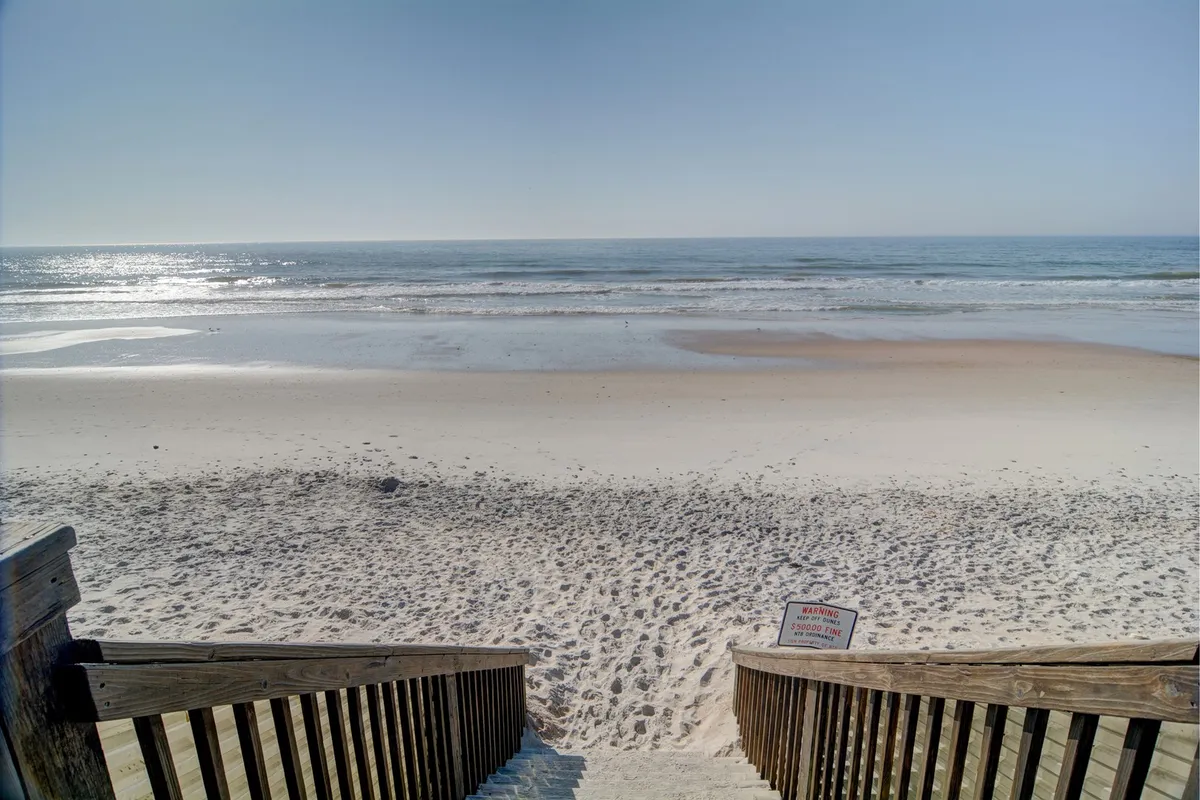 This image features stairs leading directly down to a pristine sandy beach, with gentle ocean waves rolling ashore under a bright sky. The inviting scene suggests easy, private access from the property to the secluded beach, capturing the essence of oceanfront living.