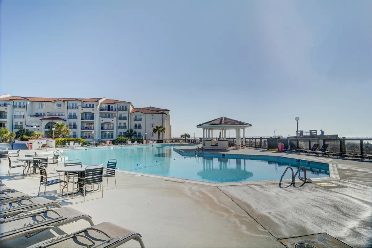 A tranquil resort swimming pool is surrounded by lounge chairs, a poolside gazebo, and the building’s inviting facade. The scene invites guests to swim, sunbathe, or socialize in a serene, sun-drenched setting with water nearby.