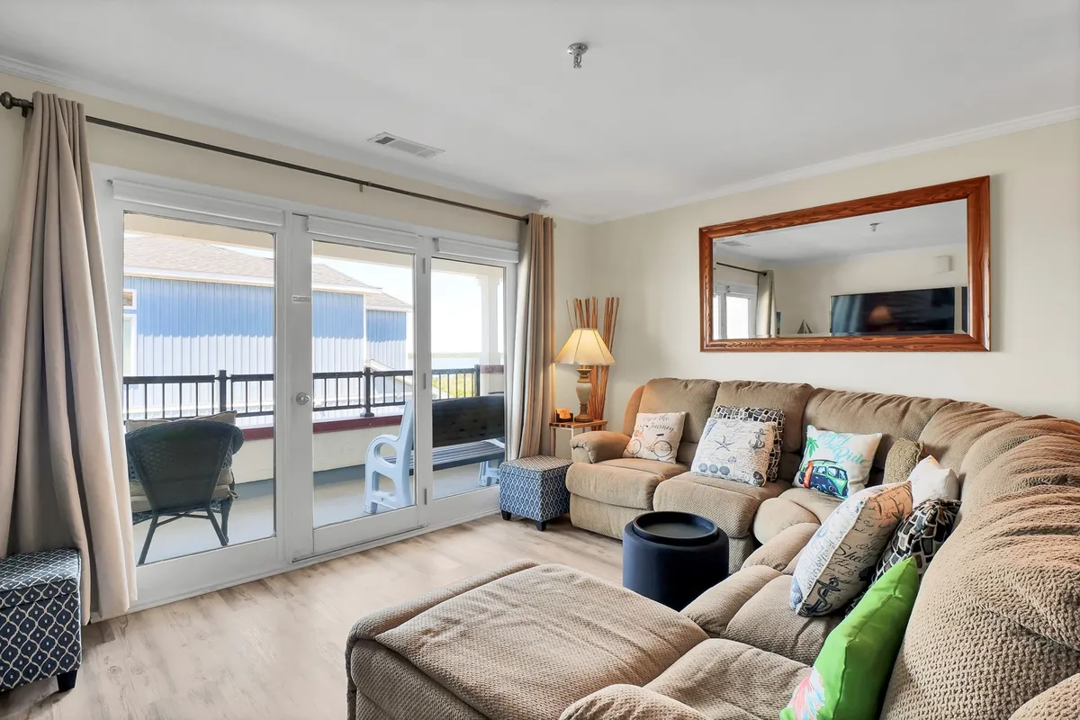 This comfortable and sunny living area features a spacious couch with colorful pillows, large windows that illuminate the room and lead to the balcony, a decorative lamp, and a framed mirror. The space is ideal for relaxing with friends or family and enjoying the abundant natural light.