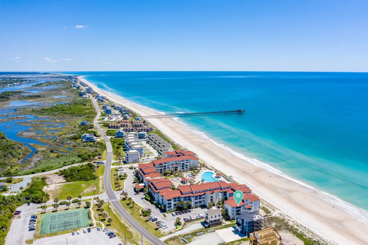 A sweeping view encompassing the sandy beach, sparkling ocean, a pier, resort buildings, and a swimming pool. There are people sunbathing, swimming, and enjoying leisurely walks along the shore. The scene is relaxed and vibrant, suggesting opportunities for beach picnics, group barbecues, and early morning outings.