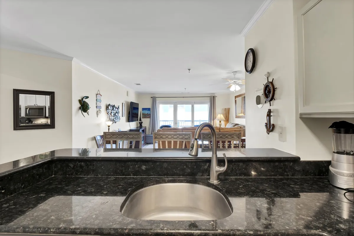 This image presents a bright, open interior where the kitchen seamlessly flows into the dining and living areas. A kitchen sink, ample counter space, and modern decor are visible, with large windows that frame a stunning view outside. The dining area is adjacent, creating a sense of togetherness and emphasizing the connection between cooking, dining, and conversation.