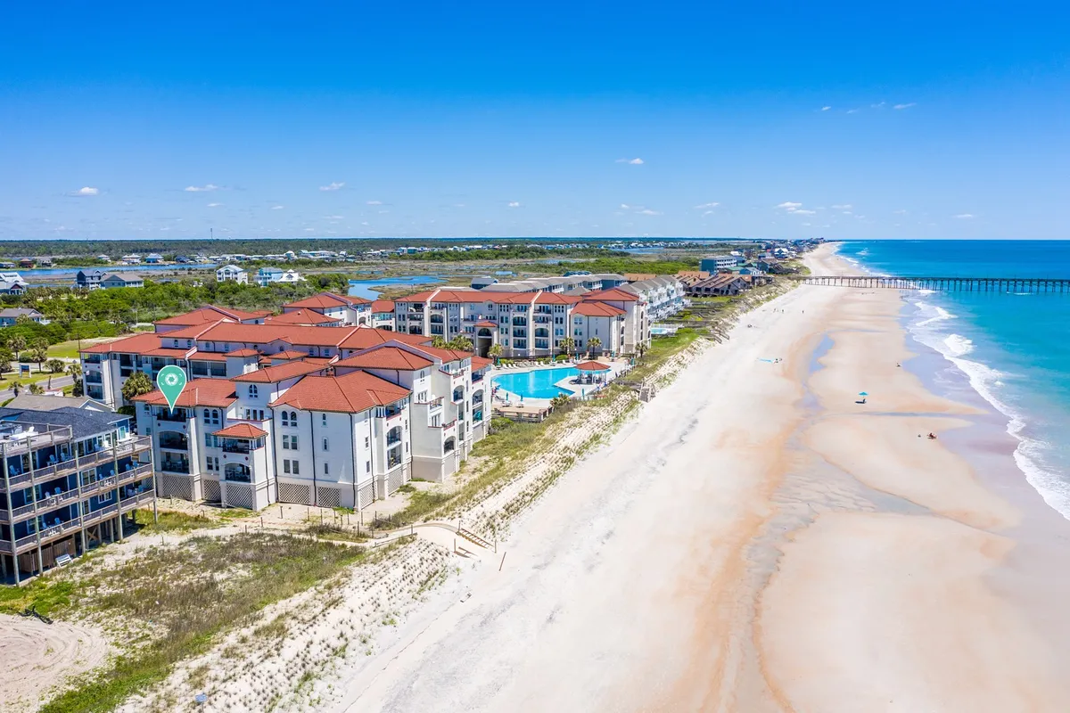 This image captures a vibrant beachfront property with a sparkling swimming pool, panoramic ocean view, the nearby pier, and an expansive sandy shoreline. The scene shows both the inviting water and the proximity to local attractions, with families able to enjoy the pool, stroll along the shore, and head out to the pier for fishing or sightseeing.
