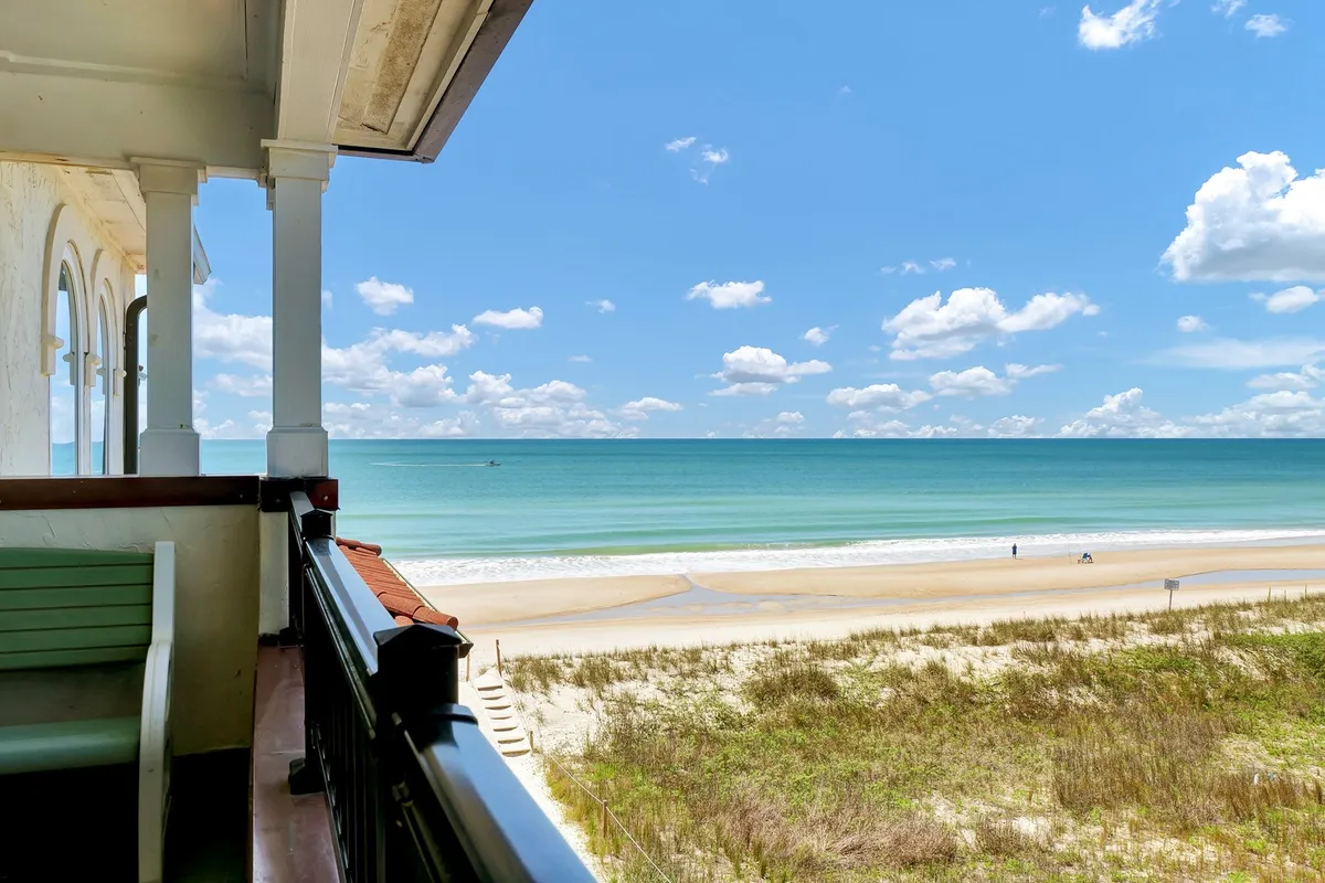 The image showcases a sweeping view of the beach and ocean from a private balcony, with soft clouds overhead. The inviting balcony is perfect for relaxing with a morning coffee or enjoying the sunset, while the expansive shoreline below invites guests to stroll, sunbathe, and soak in the tranquil seaside atmosphere.