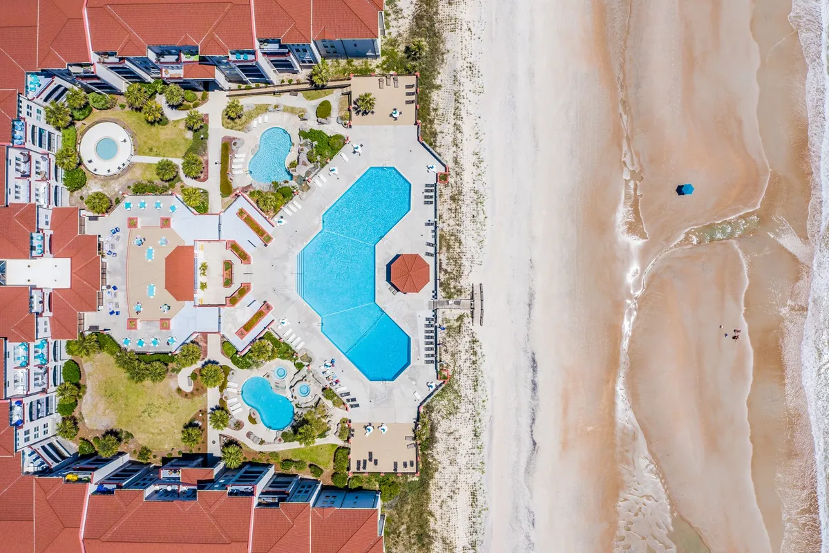 A large, inviting swimming pool is the focal point, flanked by a smaller kiddie pool and a jacuzzi. The pool area is lined with lush greenery, palm trees, and offers direct beach access, creating a true resort oasis feel. Lounge chairs are placed poolside, ideal for sunbathing or relaxing with friends and family.