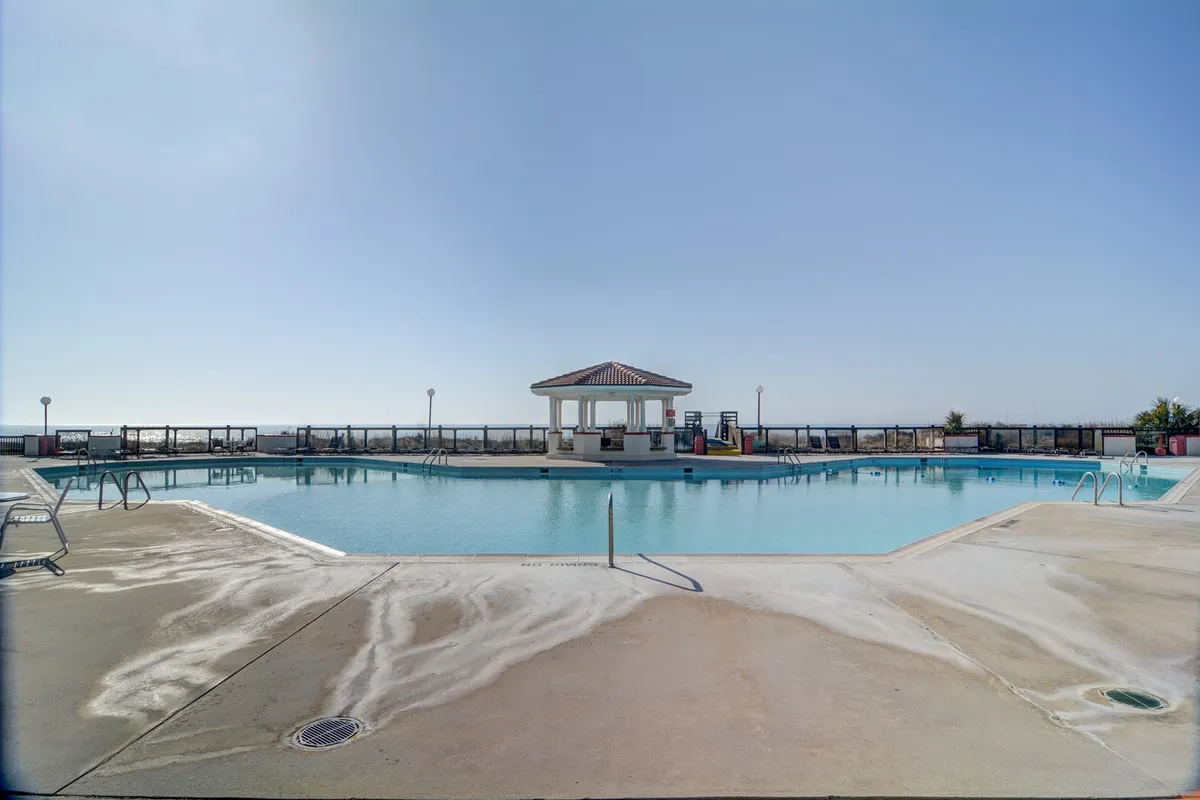 A bright exterior shot of a large swimming pool surrounded by lounge chairs and a poolside gazebo. The area is lively and inviting, perfect for both children and adults to enjoy swim time or gather for social events under the sun or in the shade.